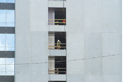 Image shows a gray building with three windows at the left and three open areas in the middle. A man in construction gear is in the middle opening.