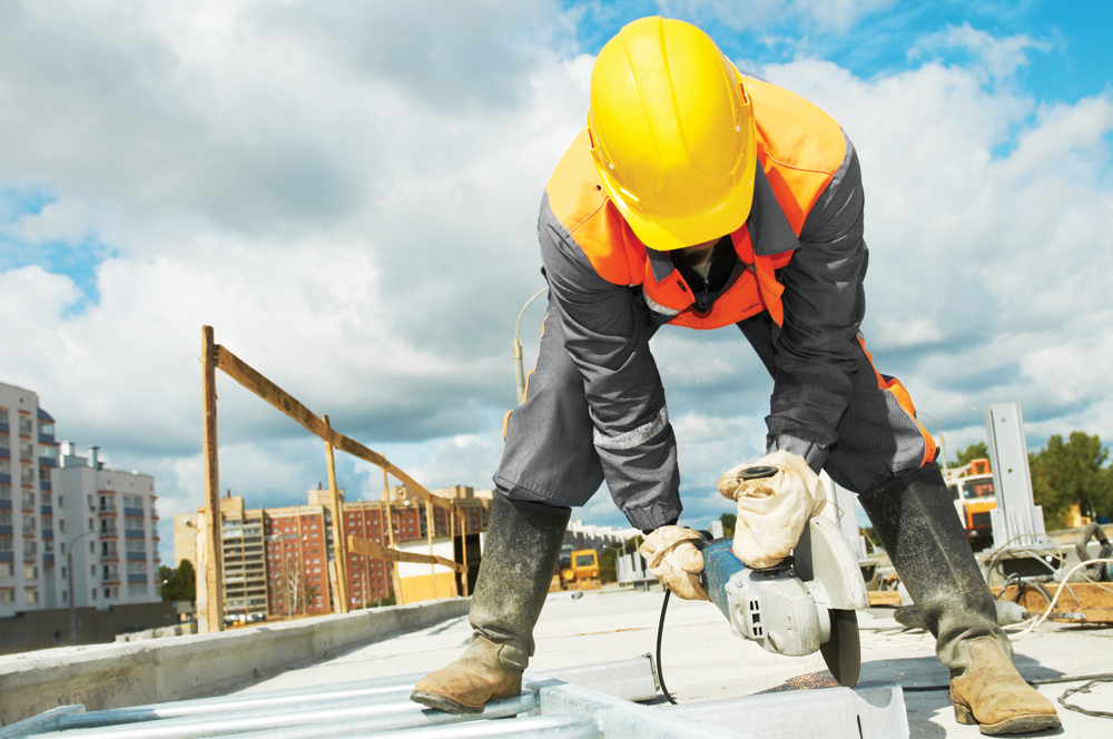 A construction worker uses a circular saw on concrete on a work site. A cityscape is behind the worker, and a blue sky with white clouds.